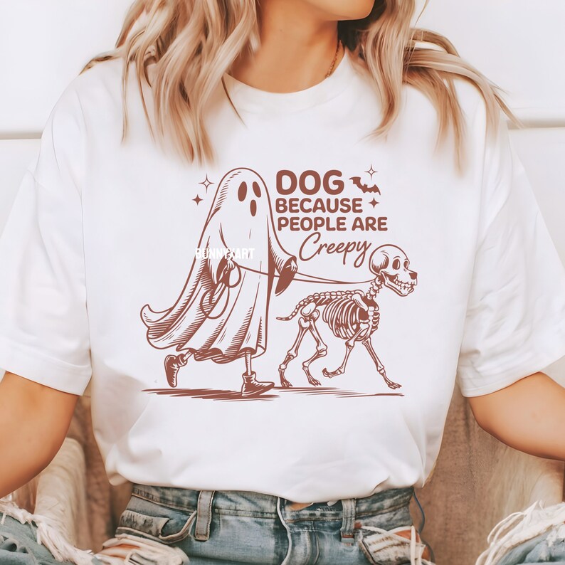 May include: White t-shirt featuring a brown graphic of a ghost walking a skeleton dog on a lead. The text says "DOG BECAUSE PEOPLE ARE CREEPY". The shirt has a crew neck and short sleeves.
