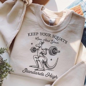 Pode incluir: Uma camisa de moletom bege com um canguru levantando pesos e o texto "KEEP YOUR SQUATS LOW AND YOUR STANDARDS HIGH".