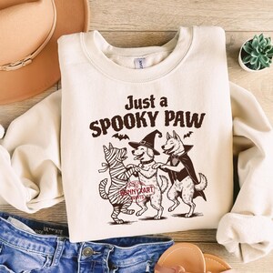 May include: Cream-colored sweatshirt with the text "Just a SPOOKY PAW" and a graphic of three dogs in Halloween costumes. One dog is wrapped in bandages, another wears a witch hat, and the third wears a cape. The sweatshirt is paired with blue jeans.