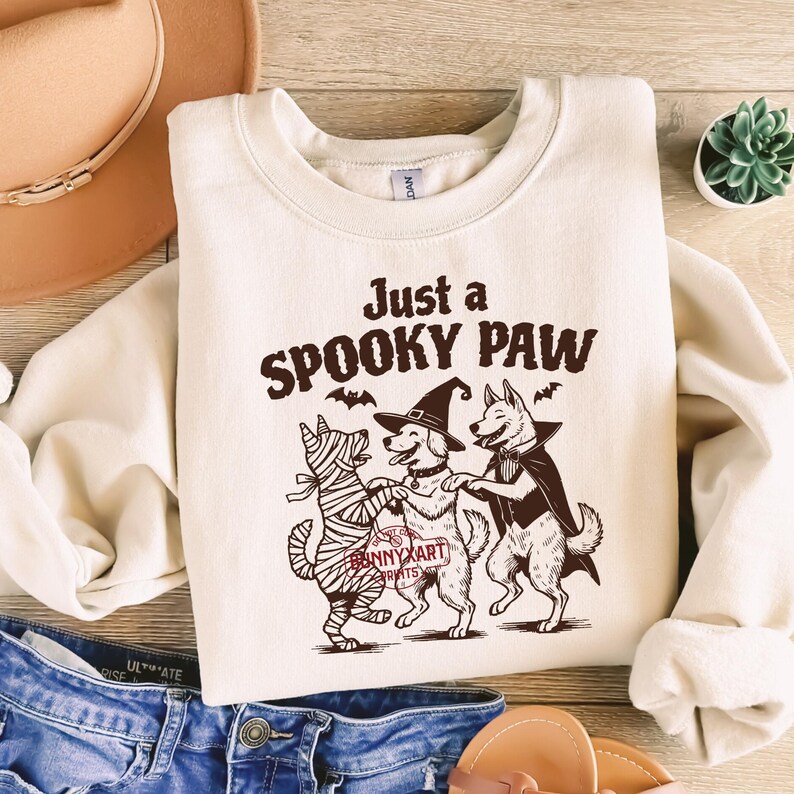 May include: Cream-colored sweatshirt with the text "Just a SPOOKY PAW" and a graphic of three dogs in Halloween costumes. One dog is wrapped in bandages, another wears a witch hat, and the third wears a cape. The sweatshirt is paired with blue jeans.