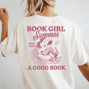 K&ouml;nnte beinhalten: Ein wei&szlig;es T-Shirt mit einem rosafarbenen Grafikdesign, das eine Meerjungfrau zeigt, die ein Buch liest. Der Text auf dem Shirt lautet "Book Girl Summer Dive Into A Good Book".