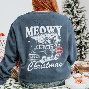 May include: A navy blue sweatshirt with a graphic featuring a truck carrying a Christmas tree and cats. The text "MEOWY Christmas" is printed in white. The design includes stars and a "Do Not Copy" watermark.