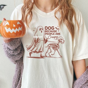 May include: A cream-coloured t-shirt with a Halloween graphic. The design shows a ghost walking a skeleton dog on a lead, with the text "DOG BECAUSE PEOPLE ARE CREEPY". The shirt has a crew neck and short sleeves.