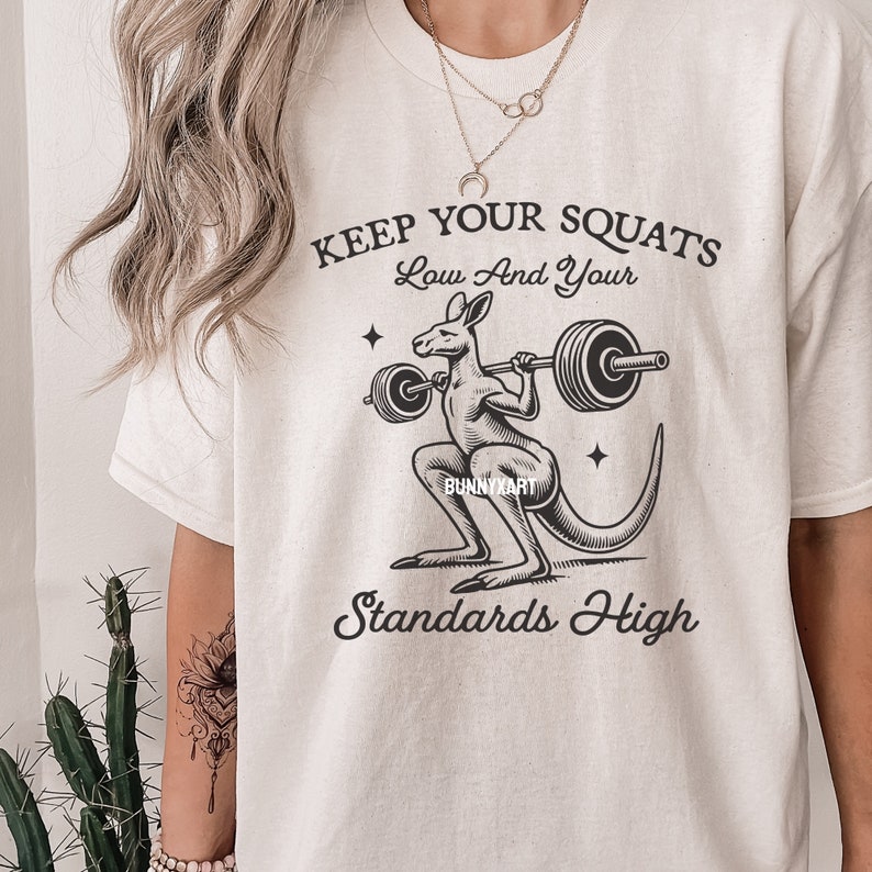 Pode incluir: Uma camiseta branca com um gr&aacute;fico preto de um canguru levantando uma barra de halteres. O texto "Keep Your Squats Low And Your Standards High" est&aacute; impresso na camiseta.