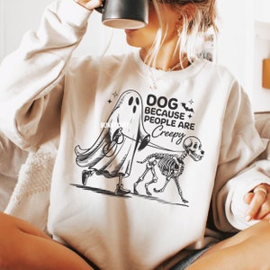 May include: Cream-coloured sweatshirt featuring a black and white graphic of a ghost walking a skeleton dog. The text reads "DOG BECAUSE PEOPLE ARE CREEPY". A spooky Halloween design.