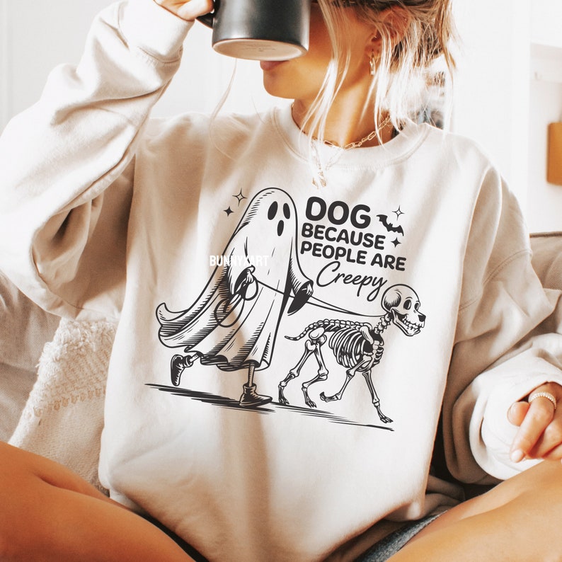 May include: Cream-coloured sweatshirt featuring a black and white graphic of a ghost walking a skeleton dog. The text reads "DOG BECAUSE PEOPLE ARE CREEPY". A spooky Halloween design.
