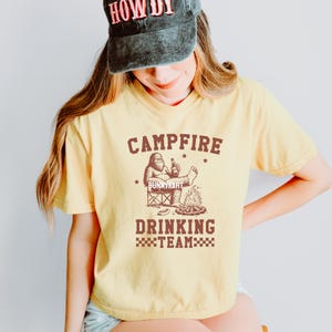 Puede incluir: Camiseta amarilla clara con un gr&aacute;fico marr&oacute;n. El gr&aacute;fico dice "Campfire Drinking Team" y presenta un personaje de Bigfoot con una botella cerca de una fogata. El texto "Bunnyxart" tambi&eacute;n es visible.