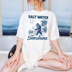 May include: Cream-colored t-shirt with a blue graphic of a Bigfoot carrying a surfboard, palm trees, and the sun. The text "SALT WATER" is above the graphic, and "and Sunshine" is below. The shirt is a relaxed fit.