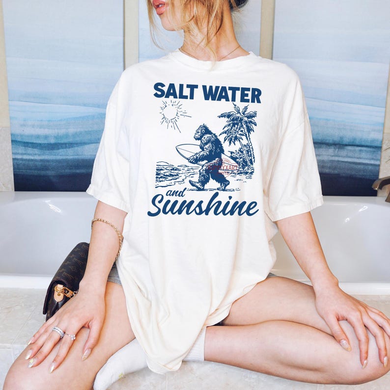 May include: Cream-colored t-shirt with a blue graphic of a Bigfoot carrying a surfboard, palm trees, and the sun. The text "SALT WATER" is above the graphic, and "and Sunshine" is below. The shirt is a relaxed fit.