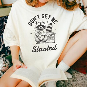 May include: A white t-shirt with a black and white illustration of a raccoon holding a loaf of bread and a jar of sourdough starter. The text "Don't get me started" is printed below the illustration.