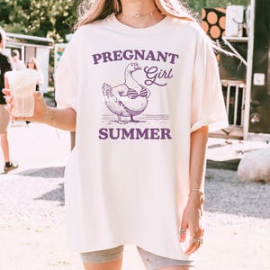 May include: A white oversized t-shirt with a purple graphic of a pregnant duck wearing a bikini and the text "PREGNANT Girl SUMMER".
