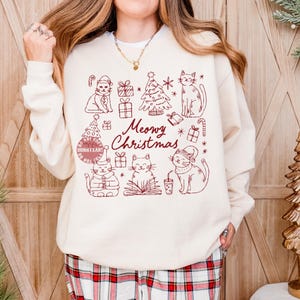 May include: Cream-colored sweatshirt with a maroon graphic featuring cats, Christmas trees, gifts, and candy canes. The text "Meowy Christmas" is written in a script font. The sweatshirt is a festive holiday design.