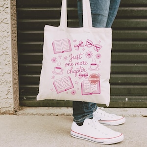 Op de afbeelding: Een witte draagtas met een roze afbeelding van boeken, strikken, brillen en koffiekopjes. De tekst "Just one more chapter" staat ook op de tas. De tas heeft twee witte handvatten.