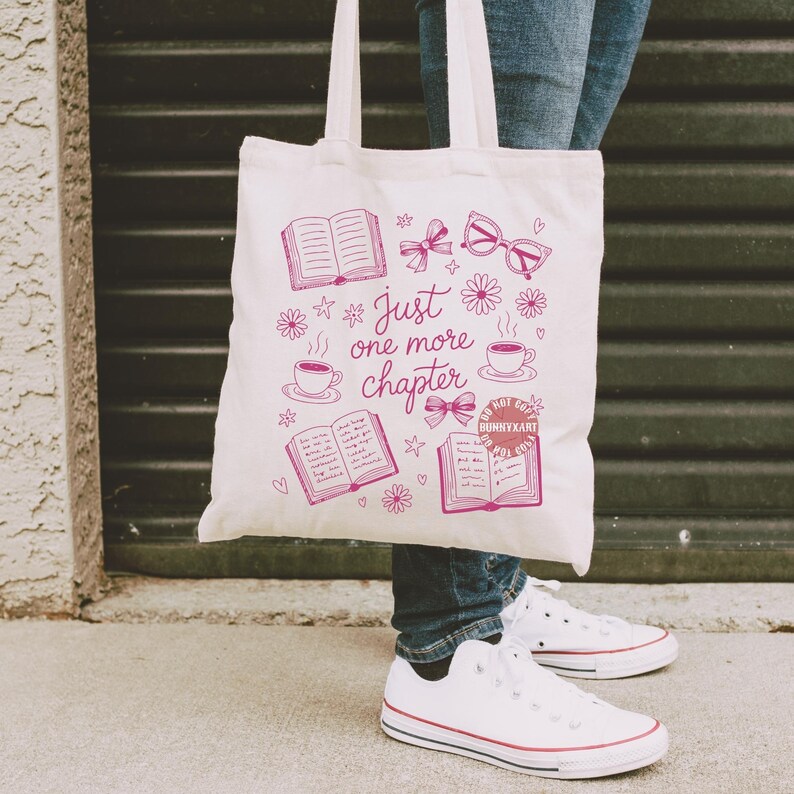 Op de afbeelding: Een witte draagtas met een roze afbeelding van boeken, strikken, brillen en koffiekopjes. De tekst "Just one more chapter" staat ook op de tas. De tas heeft twee witte handvatten.