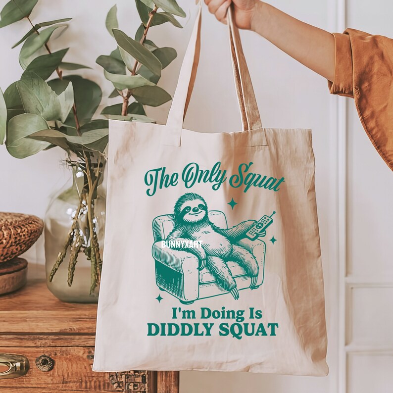 May include: A beige tote bag featuring a green sloth design. The sloth is relaxing in a chair, holding a remote control. The text on the bag reads "The Only Squat I'm Doing Is DIDDLY SQUAT".