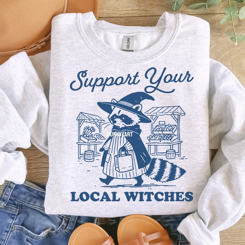 K&ouml;nnte beinhalten: Ein graues Sweatshirt mit einer blauen Grafik eines Waschb&auml;ren, der einen Hexenhut tr&auml;gt und eine Tasche h&auml;lt. Der Text "Support Your Local Witches" ist unter der Grafik gedruckt.