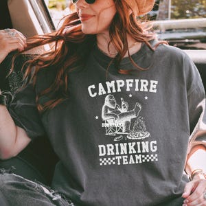 Puede incluir: Camiseta gris oscuro con las palabras "CAMPFIRE DRINKING TEAM" en blanco. Un gr&aacute;fico de una criatura parecida a Bigfoot est&aacute; sentada junto a una fogata, sosteniendo una botella. La camiseta es de estilo casual.