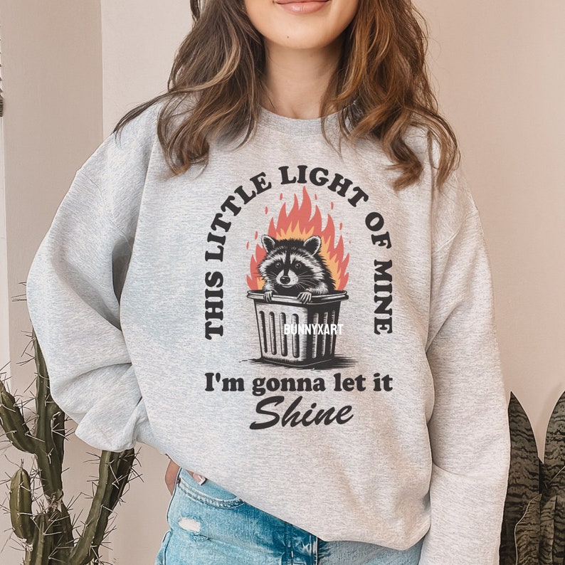 May include: Light gray sweatshirt with a graphic of a raccoon in a trash can surrounded by flames. The text reads "THIS LITTLE LIGHT OF MINE, I'm gonna let it Shine" in a circular design. The brand name "BUNNYXART" is also visible.
