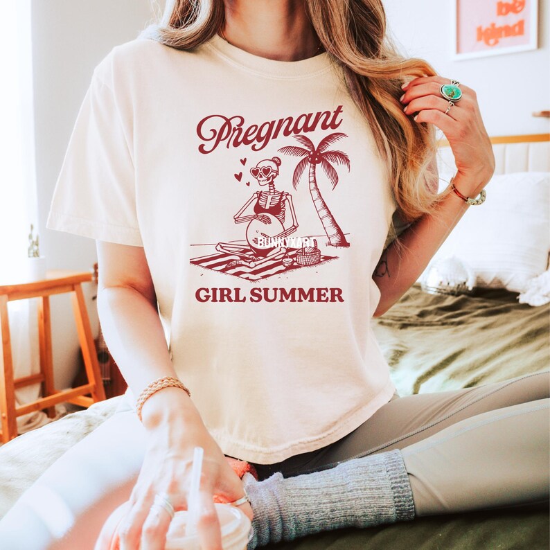 May include: A white t-shirt featuring a red graphic of a skeleton wearing sunglasses and a bikini, sitting on a beach towel with a palm tree and the text "Pregnant Girl Summer".