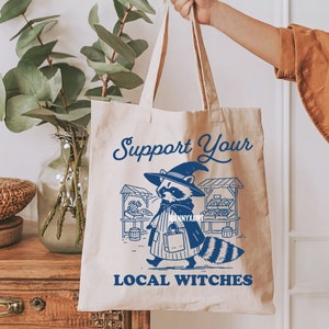 K&ouml;nnte beinhalten: Eine wei&szlig;e Canvas-Tragetasche mit blauem Text und einer Grafik eines Waschb&auml;ren, der einen Hexenhut tr&auml;gt und eine Tasche h&auml;lt. Der Text auf der Tasche lautet "Support Your Local Witches".