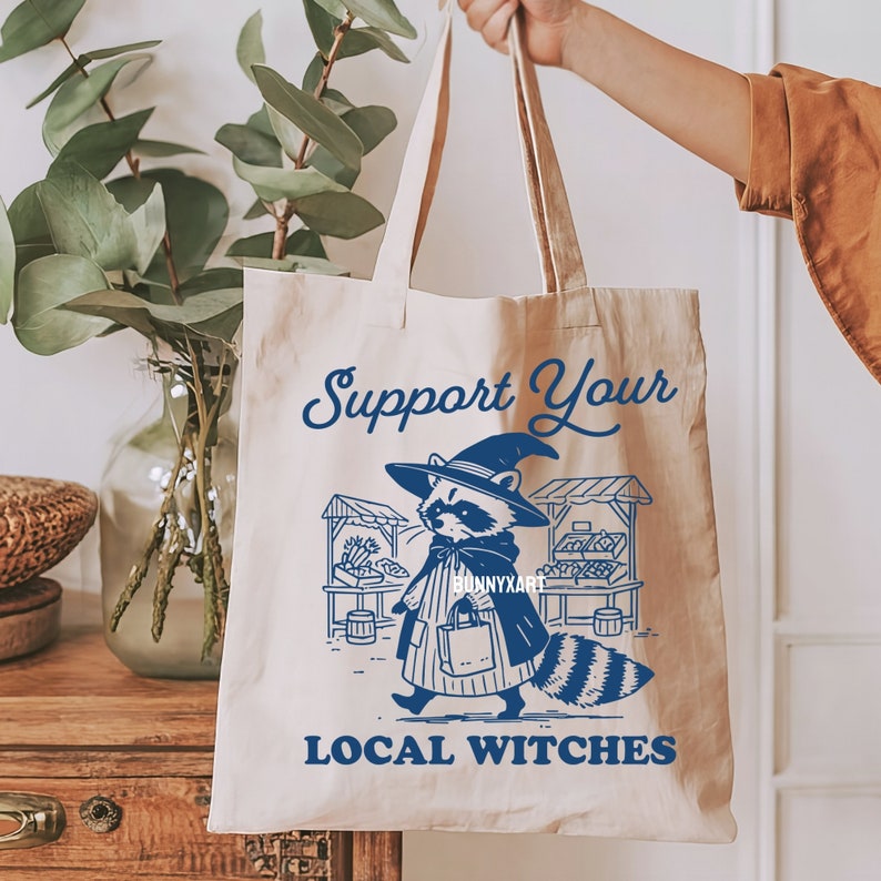 K&ouml;nnte beinhalten: Eine wei&szlig;e Canvas-Tragetasche mit blauem Text und einer Grafik eines Waschb&auml;ren, der einen Hexenhut tr&auml;gt und eine Tasche h&auml;lt. Der Text auf der Tasche lautet "Support Your Local Witches".