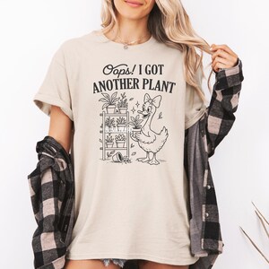 May include: Beige t-shirt with the text "Oops! I GOT ANOTHER PLANT" above a cartoon goose holding a potted plant. A plant shelf with various potted plants is to the left of the goose. The shirt is worn with a plaid shirt.