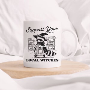 K&ouml;nnte beinhalten: Wei&szlig;e Keramiktasse mit einer Schwarzwei&szlig;illustration eines Waschb&auml;ren, der einen Hexenhut tr&auml;gt und eine Einkaufstasche h&auml;lt. Der Text "Support Your Local Witches" ist unter der Illustration aufgedruckt.