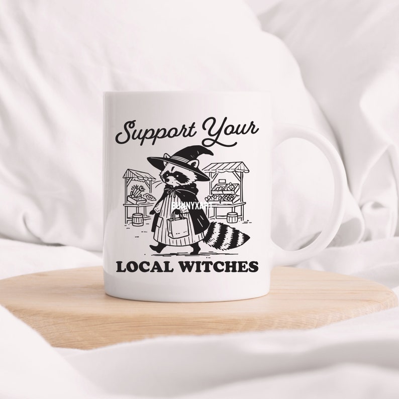 K&ouml;nnte beinhalten: Wei&szlig;e Keramiktasse mit einer Schwarzwei&szlig;illustration eines Waschb&auml;ren, der einen Hexenhut tr&auml;gt und eine Einkaufstasche h&auml;lt. Der Text "Support Your Local Witches" ist unter der Illustration aufgedruckt.