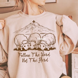May include: A beige sweatshirt with a brown graphic of a mountain with a cross on top and three sheep below. The text "Follow The Word Not The Herd" is printed below the sheep.
