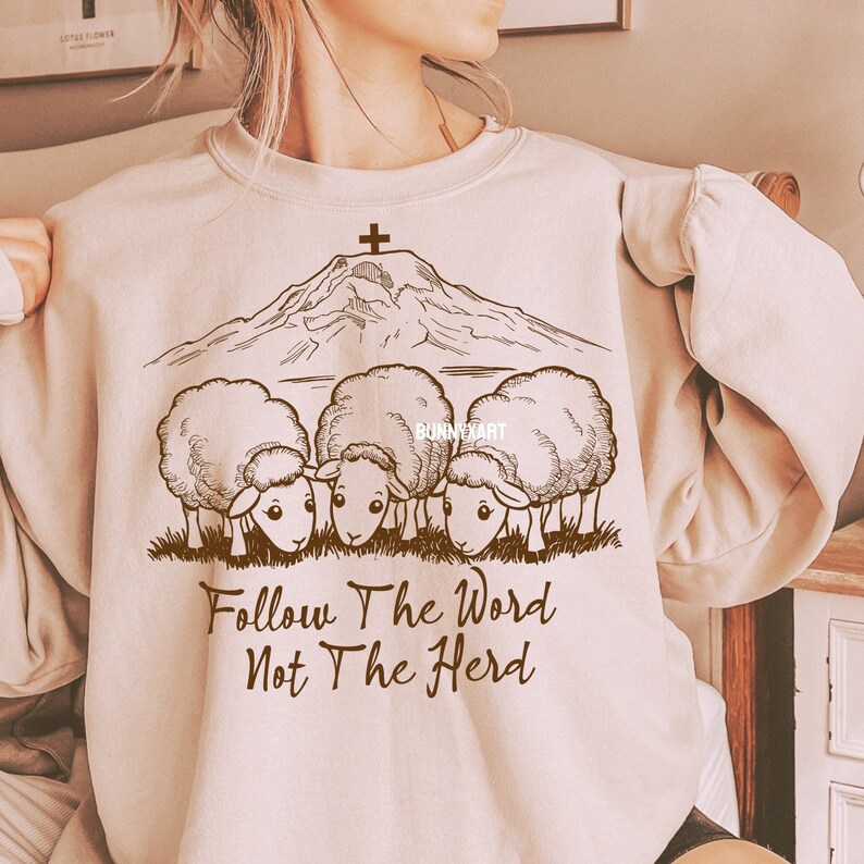 May include: A beige sweatshirt with a brown graphic of a mountain with a cross on top and three sheep below. The text "Follow The Word Not The Herd" is printed below the sheep.