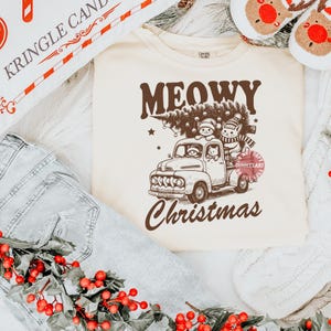 May include: Cream-colored sweatshirt with a brown graphic of a vintage truck carrying a Christmas tree and cats. The text on the shirt reads "MEOWY Christmas". The sweatshirt is laid on a white surface with a pair of jeans and a decorative pillow.
