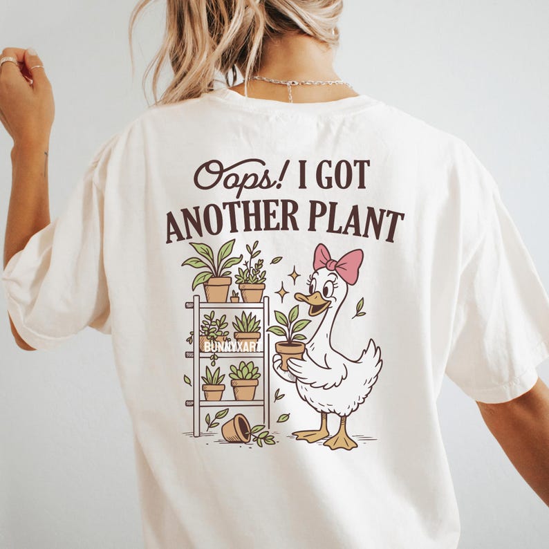 May include: Cream-colored t-shirt with the phrase "Oops! I Got Another Plant" and a cartoon duck holding a potted plant. The design includes a plant shelf with various potted plants and the text "Bunnyxart".