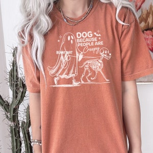 May include: Burnt orange t-shirt featuring a white graphic of a ghost walking a skeleton dog. The text on the shirt says "DOG BECAUSE PEOPLE ARE CREEPY" with the artist's name "BUNNY ART". A casual, unisex style.