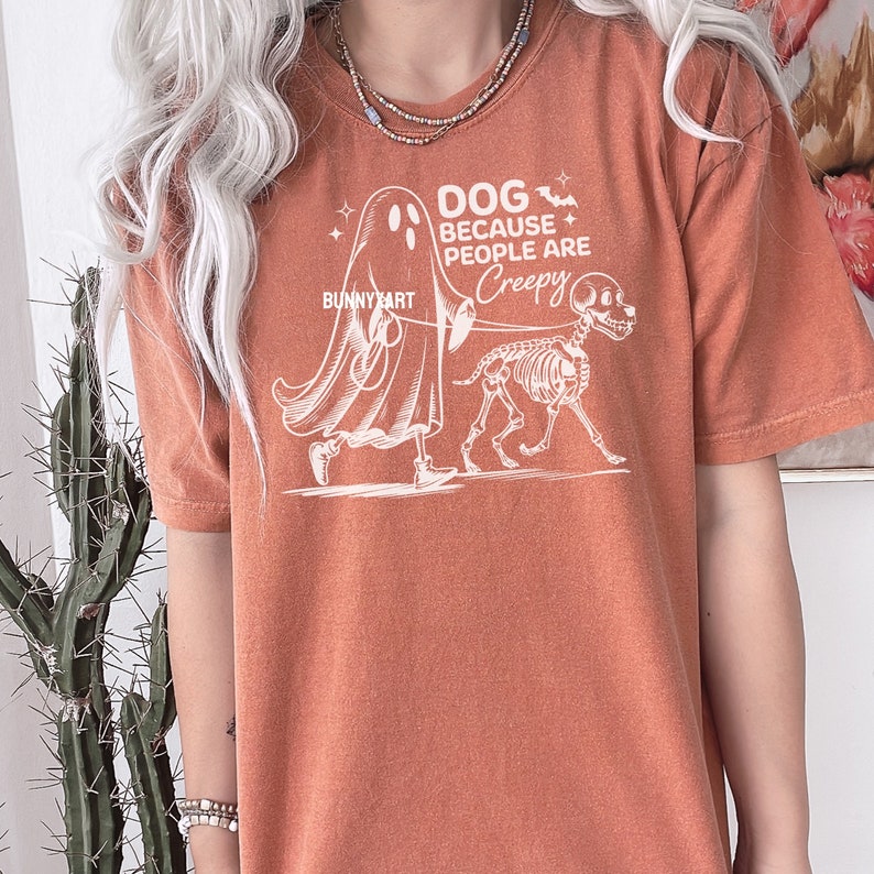 May include: Burnt orange t-shirt featuring a white graphic of a ghost walking a skeleton dog. The text on the shirt says "DOG BECAUSE PEOPLE ARE CREEPY" with the artist's name "BUNNY ART". A casual, unisex style.