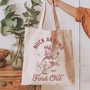 May include: A beige tote bag with a red graphic of a frog riding a bull. The text "Buck Around and Find Out" is printed in red on the bag.