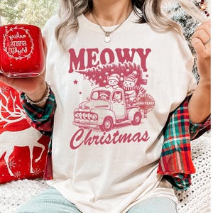 May include: Cream-colored t-shirt with a pink graphic of a vintage truck carrying a Christmas tree and cats. The text "MEOWY Christmas" is above and below the truck design. A red mug with white text is held in the image.