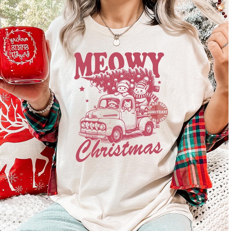 May include: Cream-colored t-shirt with a pink graphic of a vintage truck carrying a Christmas tree and cats. The text "MEOWY Christmas" is above and below the truck design. A red mug with white text is held in the image.