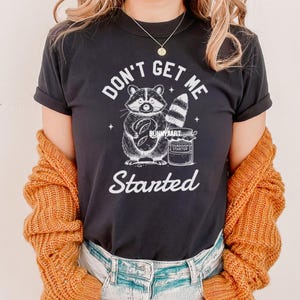 May include: Black t-shirt with a white graphic of a raccoon holding a loaf of bread and a jar of sourdough starter. The text "Don't Get Me Started" is printed below the raccoon.