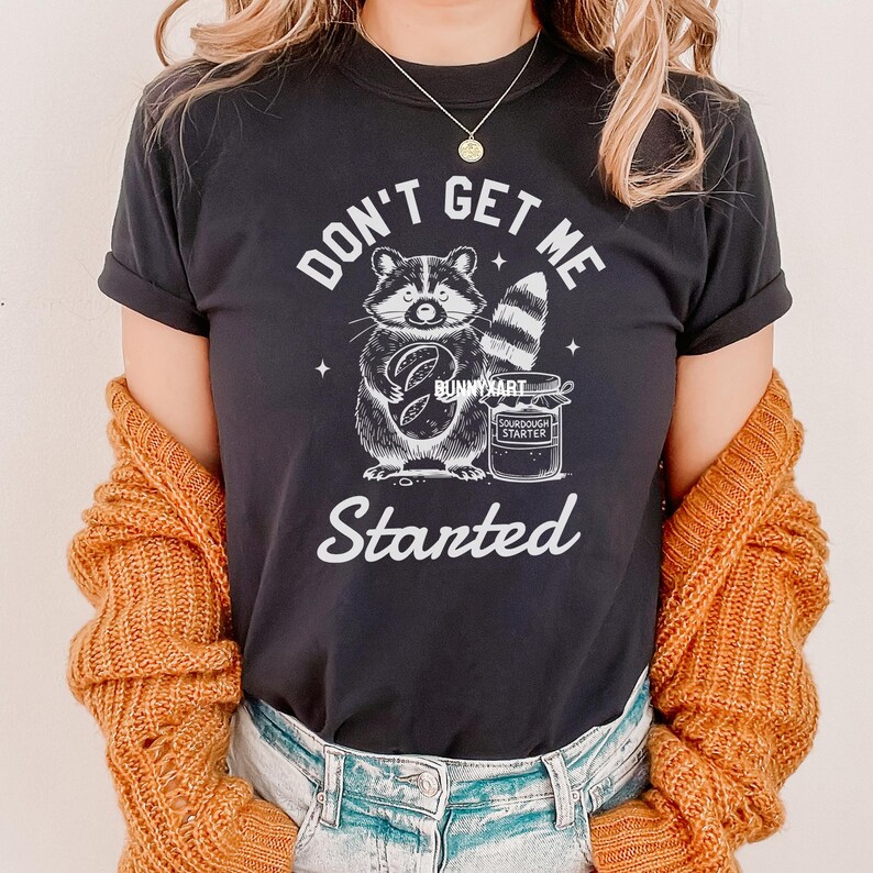 May include: Black t-shirt with a white graphic of a raccoon holding a loaf of bread and a jar of sourdough starter. The text "Don't Get Me Started" is printed below the raccoon.