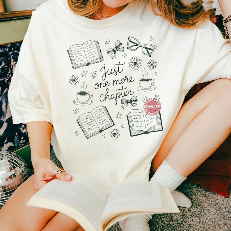 Op de afbeelding: Cr&egrave;mekleurig T-shirt met een zwart-wit grafisch ontwerp met open boeken, koffiekopjes, strikken en de tekst "Just one more chapter". De persoon die het T-shirt draagt, houdt een open boek vast.