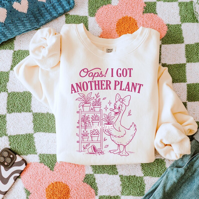 May include: Cream-colored sweatshirt with the text "Oops! I GOT ANOTHER PLANT" in magenta. A cartoon duck with a bow holds a potted plant. The design includes a plant shelf with various potted plants.