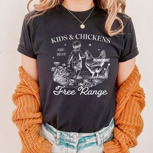 May include: Black t-shirt with a white graphic design of a child and a chicken walking in a field of flowers. The text "Kids & Chickens Are Best Free Range" is printed on the shirt.