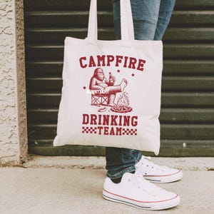 Puede incluir: Una bolsa de lona blanca con las palabras "CAMPFIRE DRINKING TEAM" en rojo. El dise&ntilde;o presenta una caricatura de Bigfoot relaj&aacute;ndose junto a una fogata, sosteniendo una cerveza. La bolsa tiene dos asas.