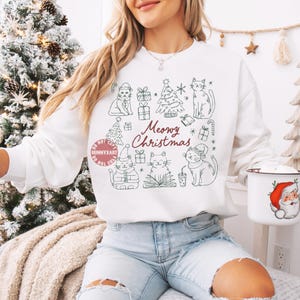 May include: White sweatshirt with a green line drawing of cats, Christmas trees, and gifts. The text "Meowy Christmas" is written in red script. A Santa mug is held in the hand.