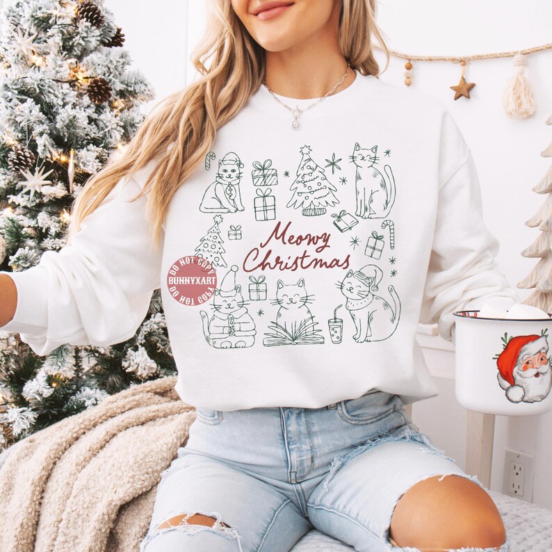 May include: White sweatshirt with a green line drawing of cats, Christmas trees, and gifts. The text "Meowy Christmas" is written in red script. A Santa mug is held in the hand.