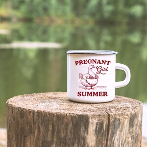 May include: White enamel camping mug with a black rim and handle. The mug has a red graphic of a pregnant duck wearing a bikini and the text "Pregnant Girl Summer".