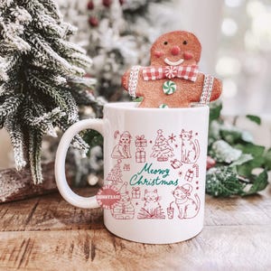 May include: White ceramic mug with a red and green cat-themed Christmas design and the words "Meowy Christmas." A gingerbread man is perched on top of the mug. The mug is on a wooden surface with a Christmas tree in the background.