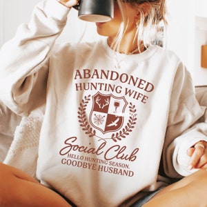 Op de afbeelding: Wit sweatshirt met bruine tekst die "Abandoned Hunting Wife Social Club Hello Hunting Season Goodbye Husband" luidt. De tekst wordt omringd door een bruin wapenschild met een hertenkop, een wijnglas, een spinnenweb en een vogel.