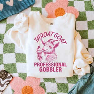May include: A cream-colored sweatshirt with a pink graphic of a goat eating an ice cream cone. The text "Throat Goat Professional Gobbler" is printed in pink on the sweatshirt.