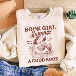 K&ouml;nnte beinhalten: Ein cremefarbener Pullover mit dem Text "Book Girl Summer" in braunen Buchstaben. Der Text "Dive Into A Good Book" ist ebenfalls in braunen Buchstaben. Eine Meerjungfrauen-Illustration befindet sich in der Mitte des Pullovers und liest ein Buch.